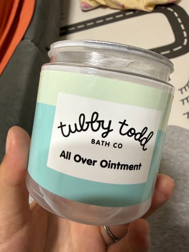 A reviewer's hand holding a clear jar of the Tubby Todd All Over Ointment.