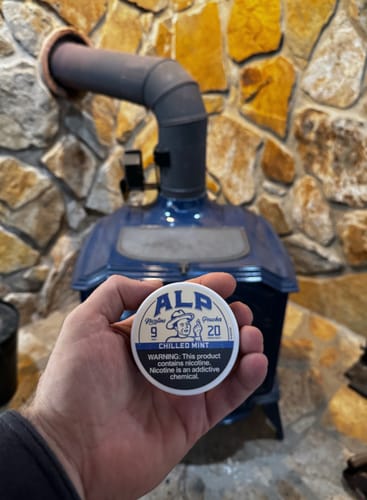 Customer holding a tin of ALP Chilled Mint Nicotine Pouches in front of a blue stove and stone wall.