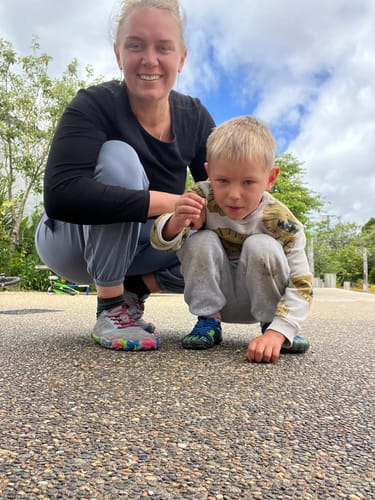 A reviewer and her son squatting outdoors. The son is wearing the blue and green Kid's Fast I - Barefoot Shoes.