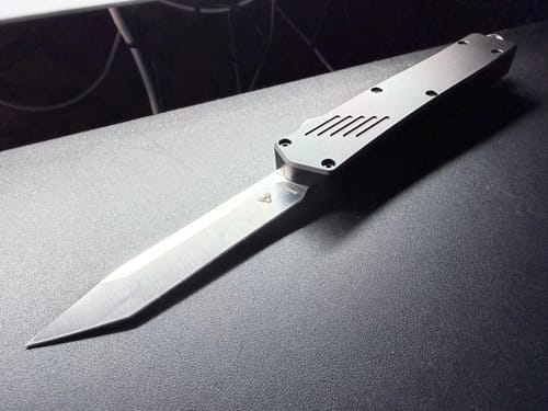 Reviewer's VT Pulse M390 OTF knife with its metallic handle and sharp M390 blade extended on a dark surface.