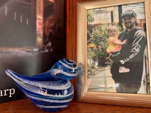 Customer's blue glass bird with cremation ash, displayed on a wooden shelf next to a framed photo.