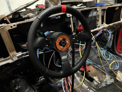 Reviewer's MPI Race Steering Wheel with red and green buttons installed inside an unfinished car with exposed wiring.