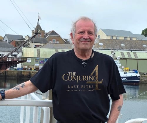 Reviewer smiling on a boat deck while wearing a black unisex t-shirt with a gold 'The Conjuring 4' graphic.