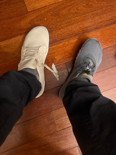 A reviewer's feet wearing two different colored Smart Ⅰ - Barefoot Shoes, one beige and one grey, on a wooden floor.