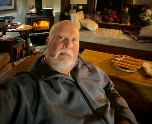 Reviewer wearing a grey zippered hoodie, sitting comfortably indoors with a fire in the background.
