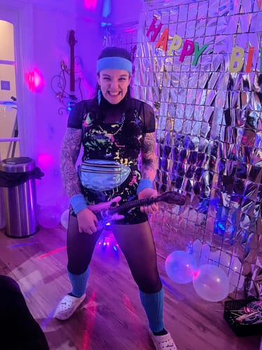 Reviewer in a colorful 80s outfit with a toy guitar, enjoying the Murder Mystery Game Kit at a party.