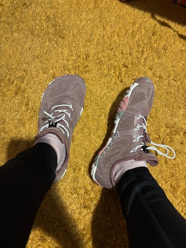 Reviewer wearing mauve Smart II Barefoot Shoes with white laces on a yellow carpet.