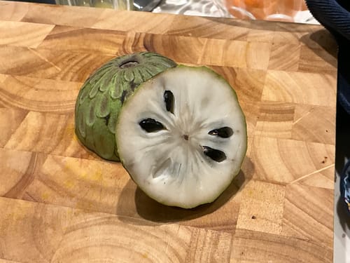 Kunde zeigt eine reife, aufgeschnittene Bio-Cherimoya mit cremigem weißen Fruchtfleisch auf einem Holzbrett.