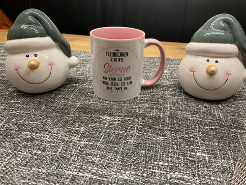 Ein Kunde zeigt die bedruckte Tasse mit rosa Henkel und Schriftzug, die auf einem Tisch zwischen zwei Schneemannfiguren steht.
