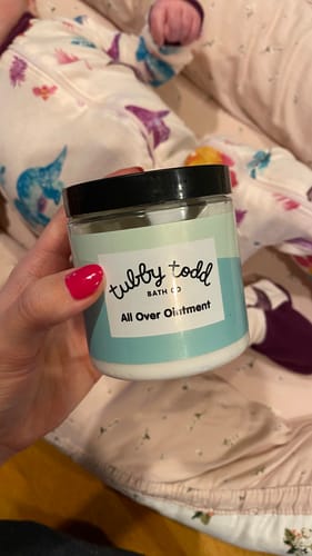 Reviewer holding a jar of All Over Ointment with a baby lying in the background.