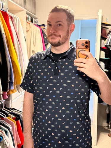 Reviewer taking a mirror selfie while wearing the patterned "All the Trimmings" polo shirt in a closet.