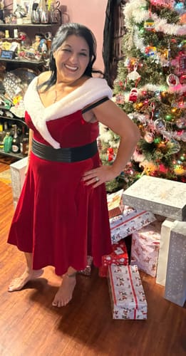 Customer wearing the red velvet Christmas dress, styled with a wide black belt while smiling by a tree.