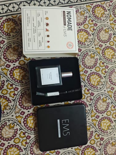 A reviewer's Nomade Eau De Parfum bottle and sampler displayed in its open black and white packaging set on a patterned cloth.