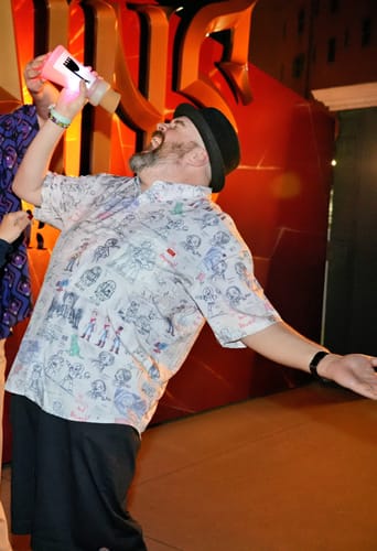 Customer in a hat wearing the "Toy Story-Board" shirt, playfully drinking from a large glowing cup indoors.