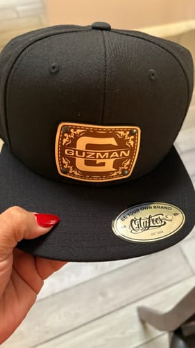 Customer holding a black Monogram Custom Hat with a leather patch personalized with the initial 'G' and the name 'GUZMAN'.