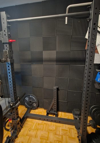 Rezensent zeigt das schwarze R7 Squat Rack, das in einem Raum mit Akustikpaneelen an der Wand aufgebaut ist.