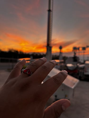 Customer's hand wearing a silver-colored promise ring with a red heart-shaped stone against a sunset.