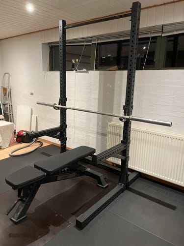 Kundenfoto des schwarzen R7 Squat Racks, aufgebaut in einem Raum mit niedriger Decke und mit einer Hantelbank.