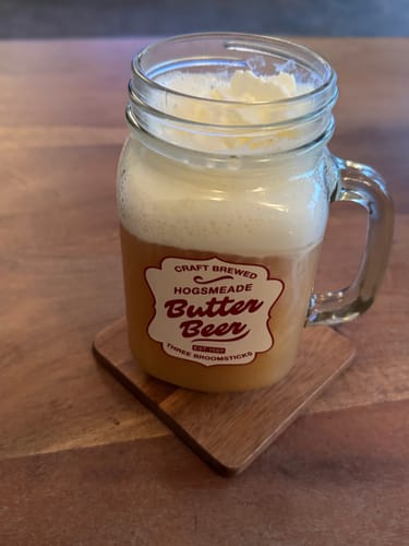 Rezensent serviert den Butterscotch Brew Tee in einem Glas mit cremiger Schaumkrone auf einem Holztisch.