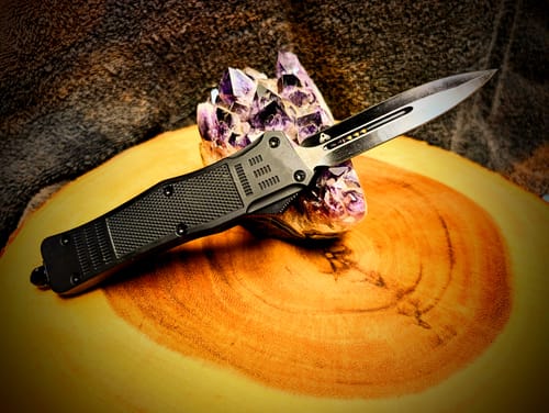 Reviewer's Black Phantom D/A OTF knife with a double-edged blade, shown resting on a purple crystal cluster.