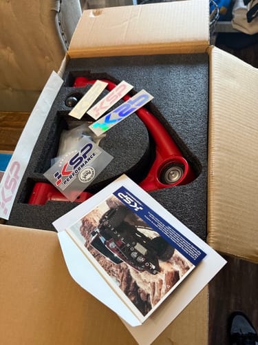 Reviewer's red Upper Control Arm in its open box with foam packaging, stickers, and an instruction booklet.