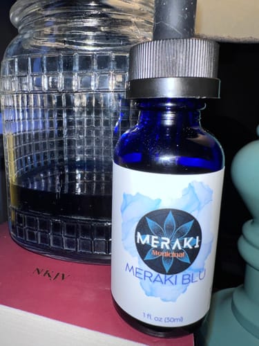 Reviewer's blue bottle of Meraki Blu sitting on a book next to a glass jar.