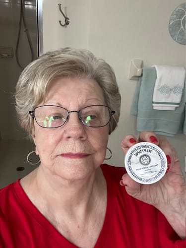 Reviewer in a red shirt holds up a white jar of Neptune Grass Fed Tallow & Honey Balm.