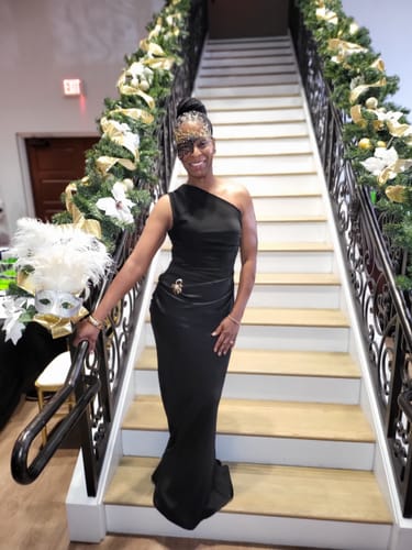 Reviewer smiling in the black Axis One Shoulder Maxi Dress, standing on a decorated staircase.