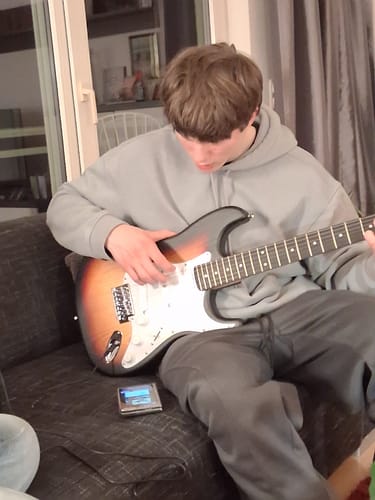 Rezensent zeigt eine Person, die auf einem Sofa eine sunburst-farbene E-Gitarre spielt.