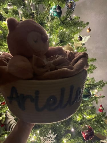 Customer holding a light-colored personalized knitted basket with the name 'Arielle', presented as a gift near a Christmas tree.