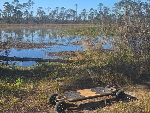 Customer's MEEPO ROVER electric skateboard parked on a dirt trail with a lake in the background.