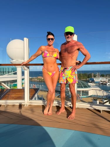 Customer and a partner in matching swimwear on a ship's deck, with the customer wearing colorful Wild Splatter Pink shorts.