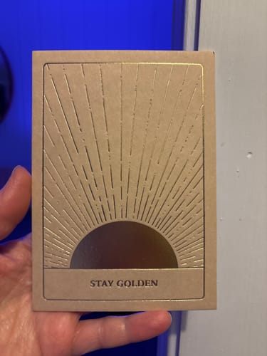 Reviewer holding the brown postcard with its gold foil sunburst design and 'STAY GOLDEN' text.