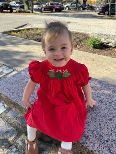Customer showing the red Christmas Tree Smocked Corduroy Dress on a smiling child sitting outdoors.