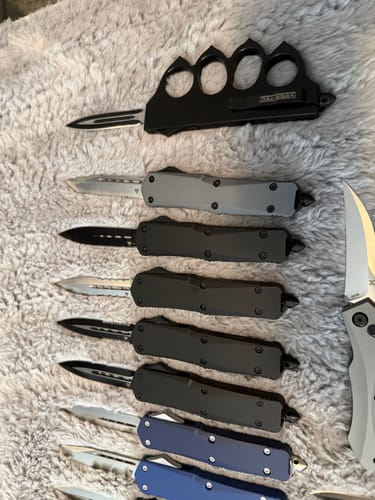Customer's black VT 1918 Trench Knuckle OTF Knife with blade extended, shown above other knives on a soft surface.