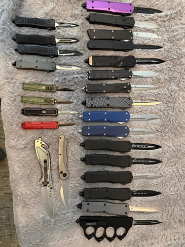Reviewer's large collection of various OTF knives, including the VT Apex D2, on a gray fuzzy surface.