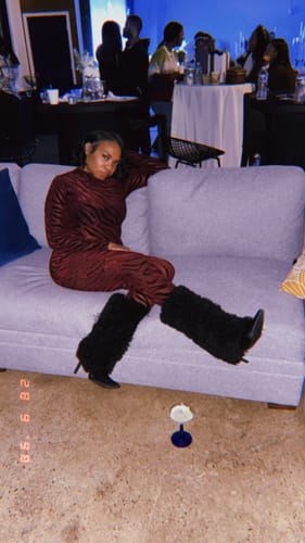 Reviewer lounging on a couch wearing the red patterned Burnout Closed Toe Jumpsuit with black furry boots.