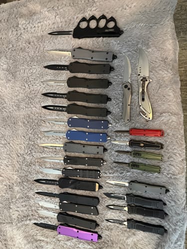 Reviewer's large collection of various OTF knives, including the Mini OTF, displayed on a fluffy gray surface.