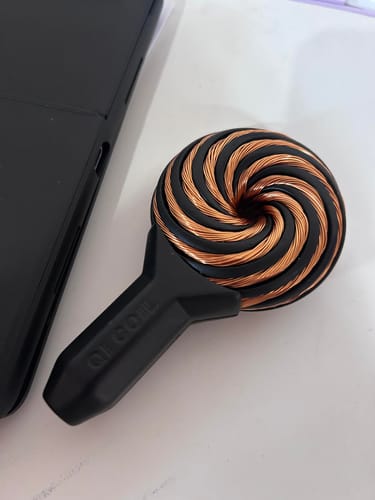 Reviewer's Qi Coil™ Mini System with its copper-wound head, resting on a surface next to a smartphone.