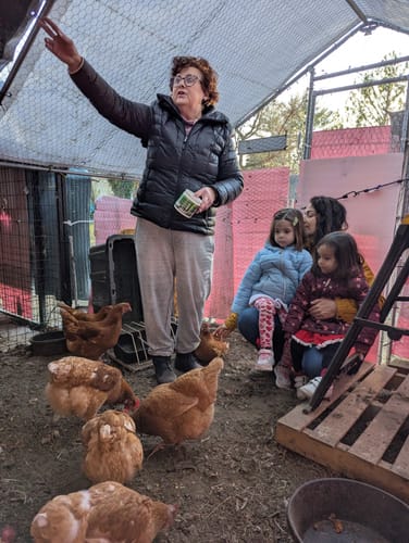 Customer in a chicken coop holding the Daily Deworm + Gut Support container, with several chickens and her family nearby.