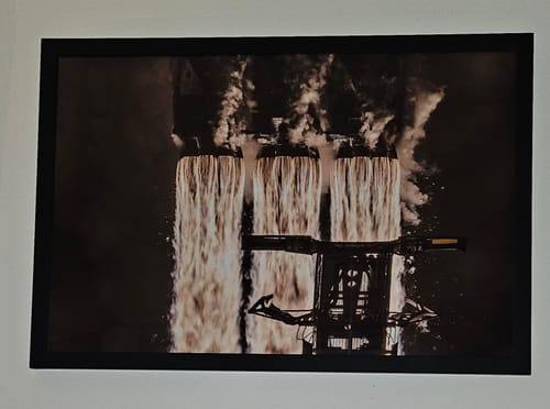 Reviewer's black-framed 'Falcon Heavy - 27 Merlins' print hung on a wall, showing the rocket's engines firing at night.