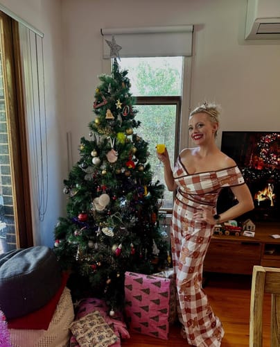 Reviewer smiling in the festive off-shoulder Gingerbread Jumpsuit, standing by a Christmas tree.