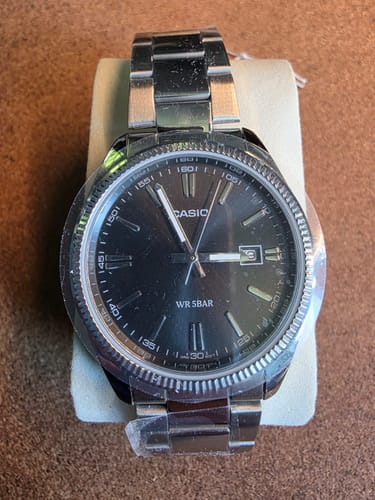 Reseñador mostrando el Reloj Casio Análogo con esfera negra y correa de acero, sobre un soporte.