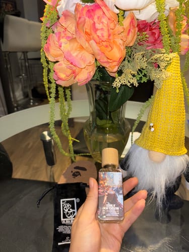 Reviewer holding a clear bottle of Evening Of Cacao Temptation perfume in their hand with a vase of flowers in the background.