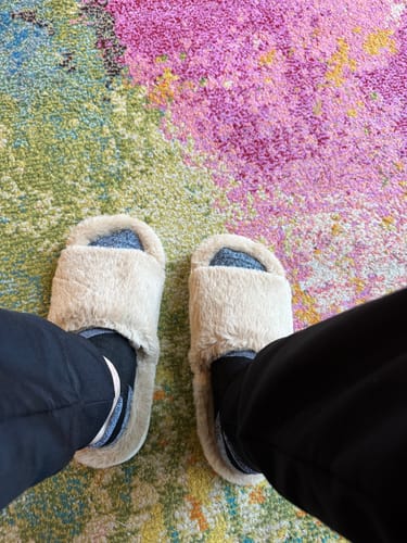 A top-down view of a customer wearing fluffy, beige slides on a colorful rug.