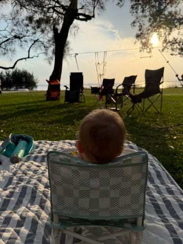 Customer's baby sits in the Parker baby chair v2 on a picnic blanket outdoors, with a sunset view over water.