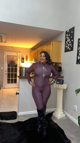 Customer wearing the purple Burnout Closed Toe Jumpsuit with tall black boots and necklaces indoors.