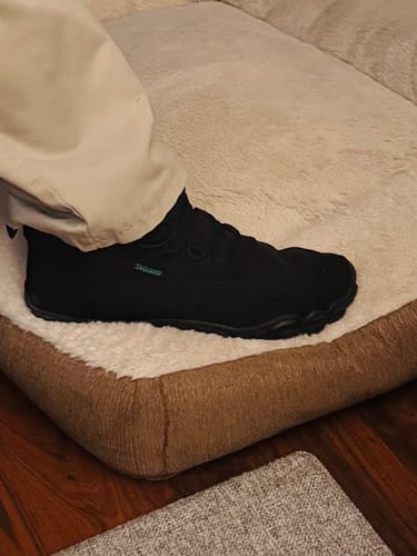 Customer wearing the black wide-toe shoes on a fluffy beige rug.