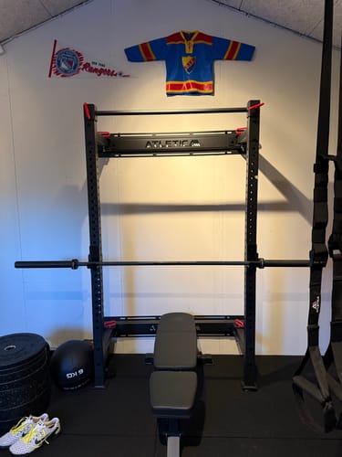 Rezensent zeigt das montierte R7 Wall Rack in einem Heim-Fitnessstudio, mit einer Hantelstange und einer Bank.