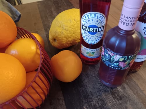 Rezensent zeigt Früchte aus der Überraschungs-Box: Orangen und eine Zitrone neben Aperitif-Flaschen für Cocktails.